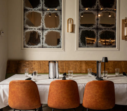 Cozy modern restaurant dining table set with white tablecloth, tan leather chairs, menus and glassware against a wooden banquette and frosted grid mirrors.