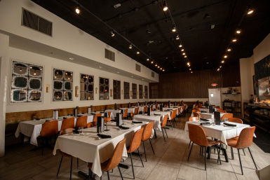 Spacious urban restaurant interior with rows of white-tablecloth tables, tan leather chairs, industrial black ceiling and warm track lighting, set and ready for service.