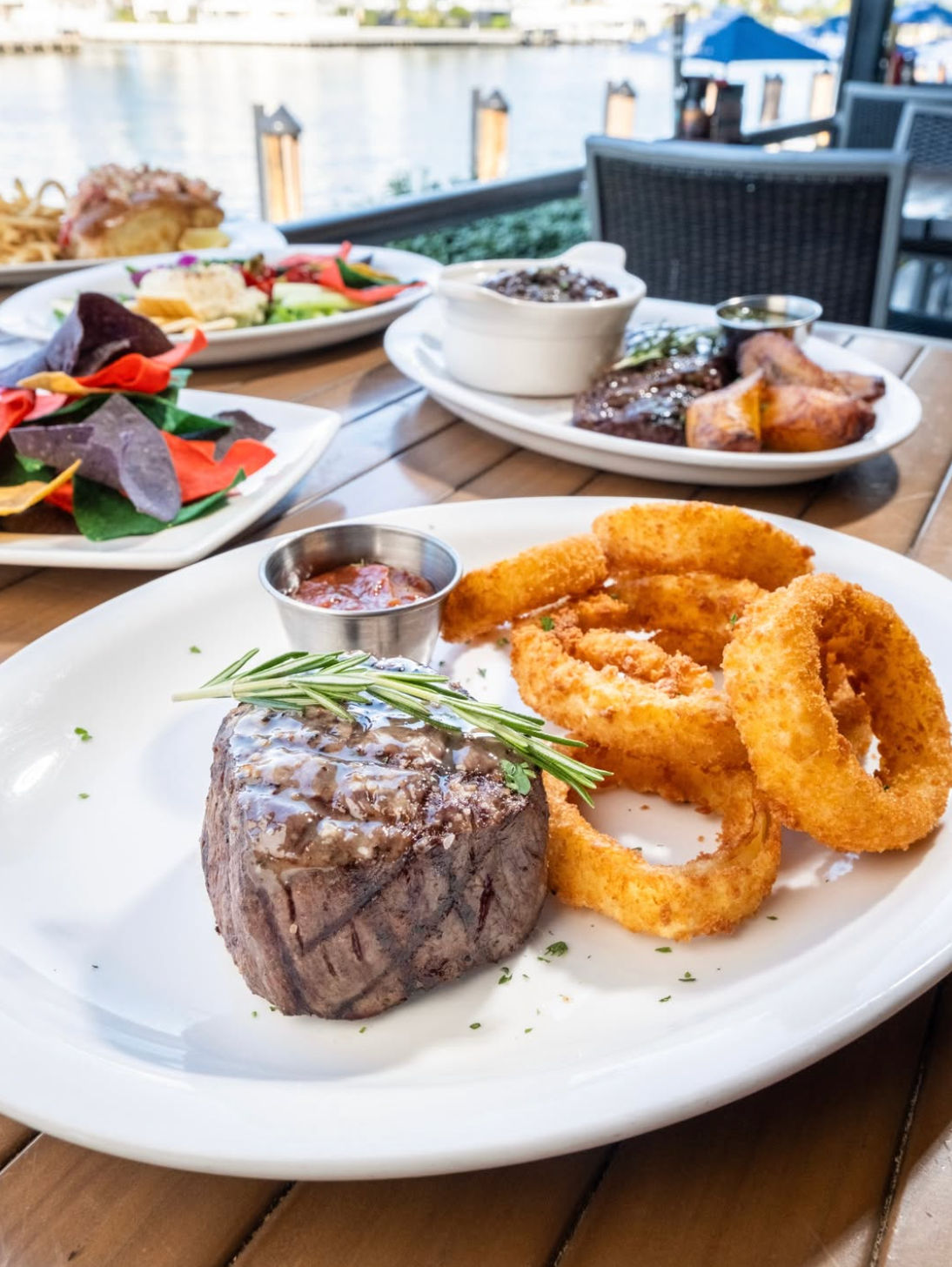 Juicy filet mignon topped with rosemary and dipping sauce alongside crispy onion rings on a waterfront restaurant patio by the marina, colorful chips and sides in background.