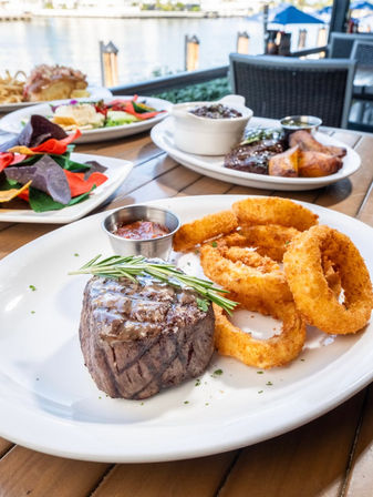 Juicy filet mignon topped with rosemary and dipping sauce alongside crispy onion rings on a waterfront restaurant patio by the marina, colorful chips and sides in background.