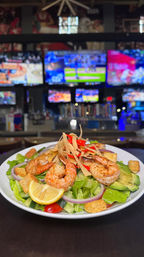 Grilled shrimp salad topped with tortilla strips, avocado, croutons and lemon wedge on a bar counter with multiple sports TVs blurred in the background.