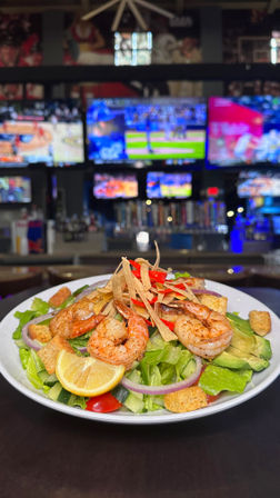 Grilled shrimp salad topped with tortilla strips, avocado, croutons and lemon wedge on a bar counter with multiple sports TVs blurred in the background.