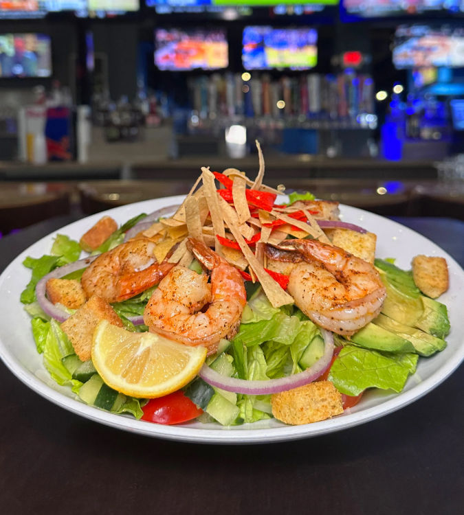 Grilled shrimp salad with avocado, romaine, red onion, cherry tomatoes, lemon wedge, croutons and crispy tortilla strips on a plate with blurred sports-bar TVs in the background.