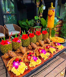 Colorful tropical brunch spread on an outdoor patio: hollowed pineapples with flowers and straws, tall citrus dispenser, waffles topped with scrambled eggs, berries and edible flowers, crispy chicken wings and a plate of orange slices amid lush plants and wicker seating.