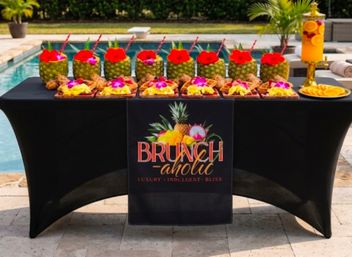 Poolside tropical brunch table with hollowed pineapple cocktails topped with flowers, fruit-topped waffles, and a citrus drink dispenser on a black tablecloth.