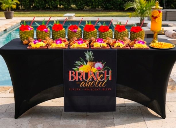 Poolside tropical brunch table with hollowed pineapple cocktails topped with flowers, fruit-topped waffles, and a citrus drink dispenser on a black tablecloth.