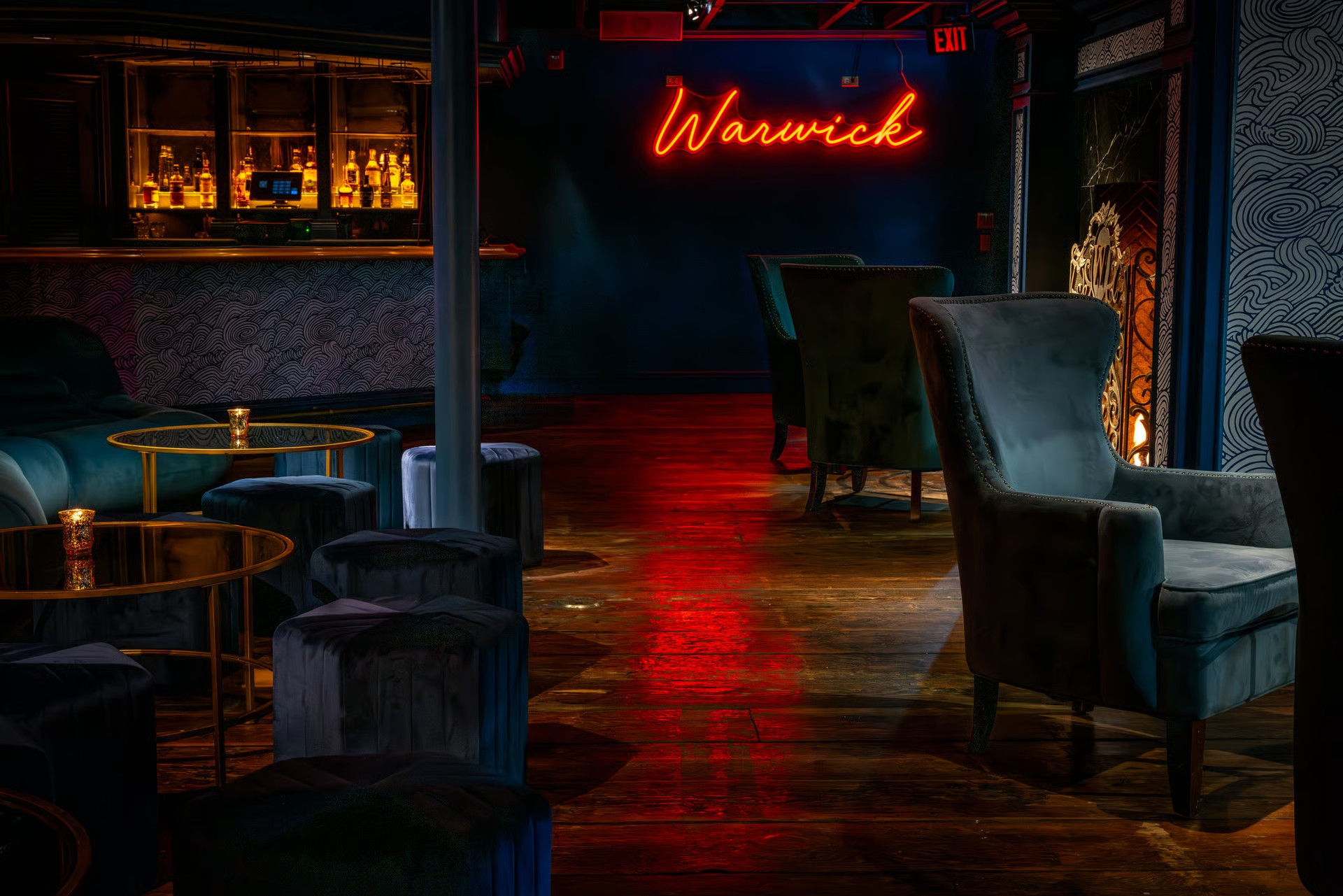 Moody upscale cocktail lounge interior with velvet armchairs, brass side tables, candles, backlit liquor shelves and a red neon sign casting a glowing reflection on polished wood floors.