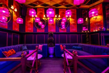 Moody neon-pink cocktail lounge with plush velvet banquettes, fringe pendant lamps, round mirrors, red crane-pattern wallpaper, marble tables and gold accents — intimate VIP seating for urban nightlife.