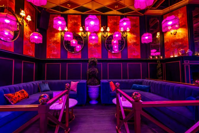 Moody neon-pink cocktail lounge with plush velvet banquettes, fringe pendant lamps, round mirrors, red crane-pattern wallpaper, marble tables and gold accents — intimate VIP seating for urban nightlife.