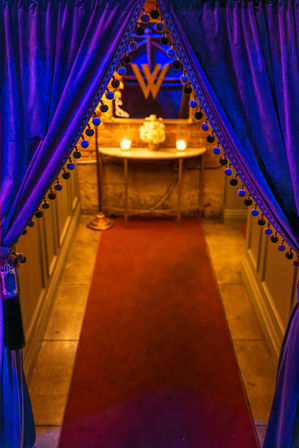 Purple velvet curtains with pom-pom trim parting to a candlelit, red-carpeted hallway leading to a small console table with flowers and warm golden lighting—cozy upscale entryway vibe.