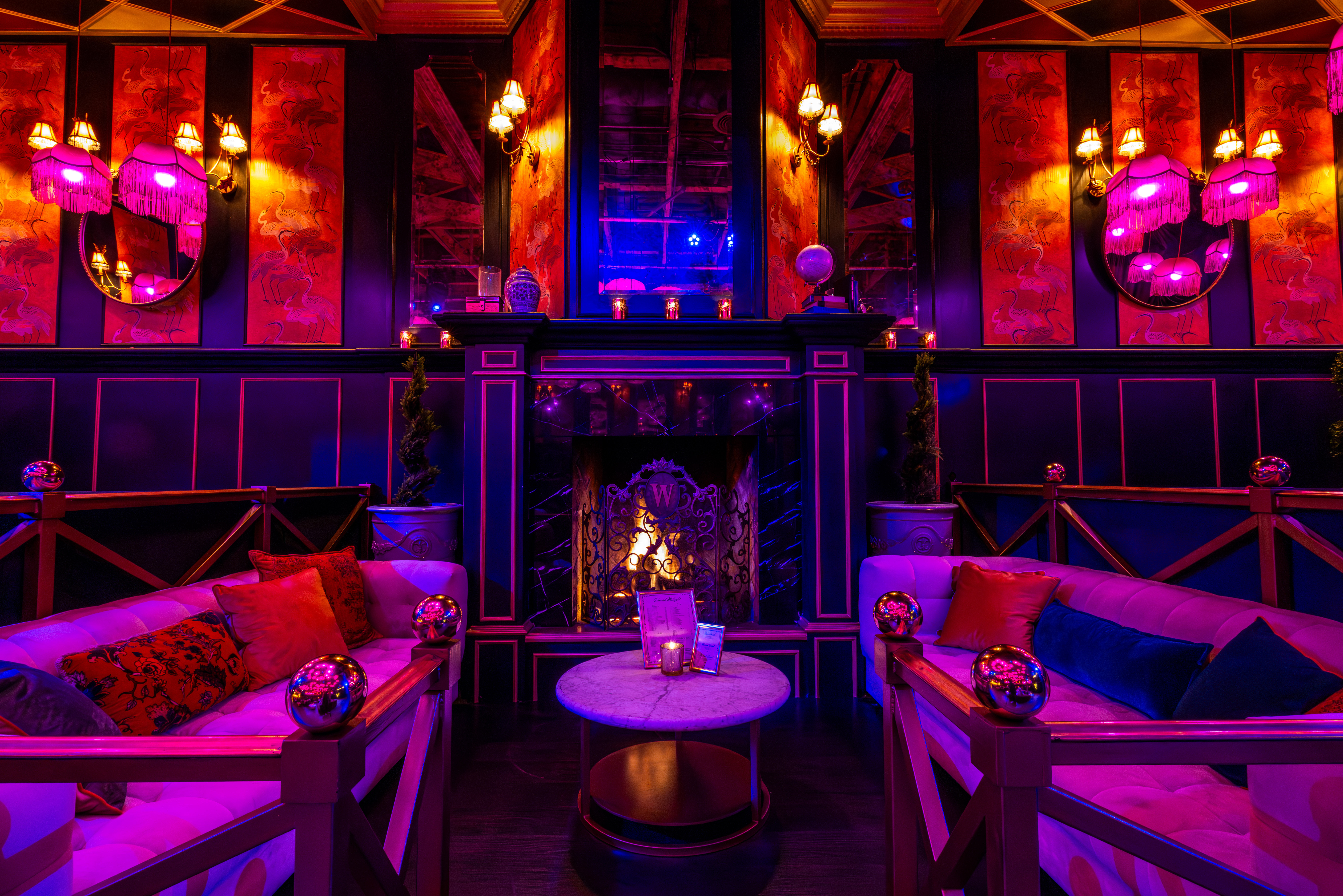 Glam vintage lounge interior with plush velvet sofas, marble coffee table, glowing fireplace and pink‑purple ambient lighting, fringe lamps and decorative mirrors