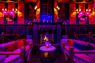 Glam vintage lounge interior with plush velvet sofas, marble coffee table, glowing fireplace and pink‑purple ambient lighting, fringe lamps and decorative mirrors