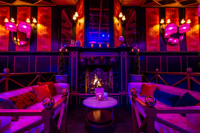 Glam vintage lounge interior with plush velvet sofas, marble coffee table, glowing fireplace and pink‑purple ambient lighting, fringe lamps and decorative mirrors