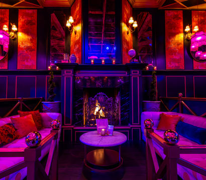 Glam vintage lounge interior with plush velvet sofas, marble coffee table, glowing fireplace and pink‑purple ambient lighting, fringe lamps and decorative mirrors