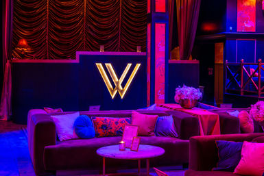 Intimate upscale lounge interior with plush velvet sofas, colorful throw pillows, marble coffee table, floral centerpieces and a glowing gold 'W' logo under moody purple-pink neon lighting