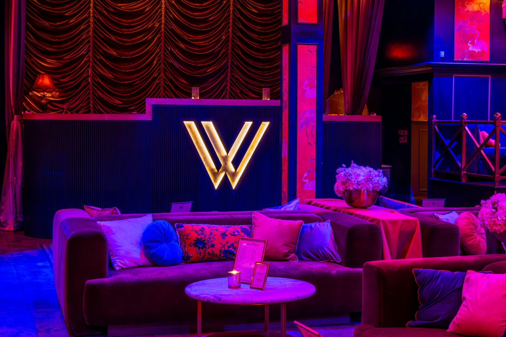 Intimate upscale lounge interior with plush velvet sofas, colorful throw pillows, marble coffee table, floral centerpieces and a glowing gold 'W' logo under moody purple-pink neon lighting