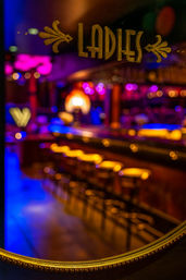 Gold-painted 'LADIES' sign on glass with blurred neon-lit cocktail bar interior, glowing amber bar stools and magenta-blue bokeh — vibrant nightlife scene