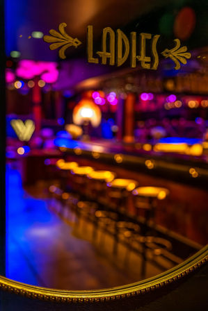 Gold-painted 'LADIES' sign on glass with blurred neon-lit cocktail bar interior, glowing amber bar stools and magenta-blue bokeh — vibrant nightlife scene