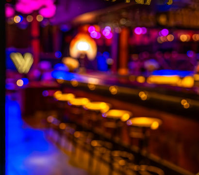 Gold-painted 'LADIES' sign on glass with blurred neon-lit cocktail bar interior, glowing amber bar stools and magenta-blue bokeh — vibrant nightlife scene