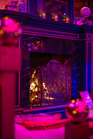 Neon-pink and purple-lit luxury lounge fireplace with black marble mantel, candles and ornate wrought-iron screen and warm crackling flames.