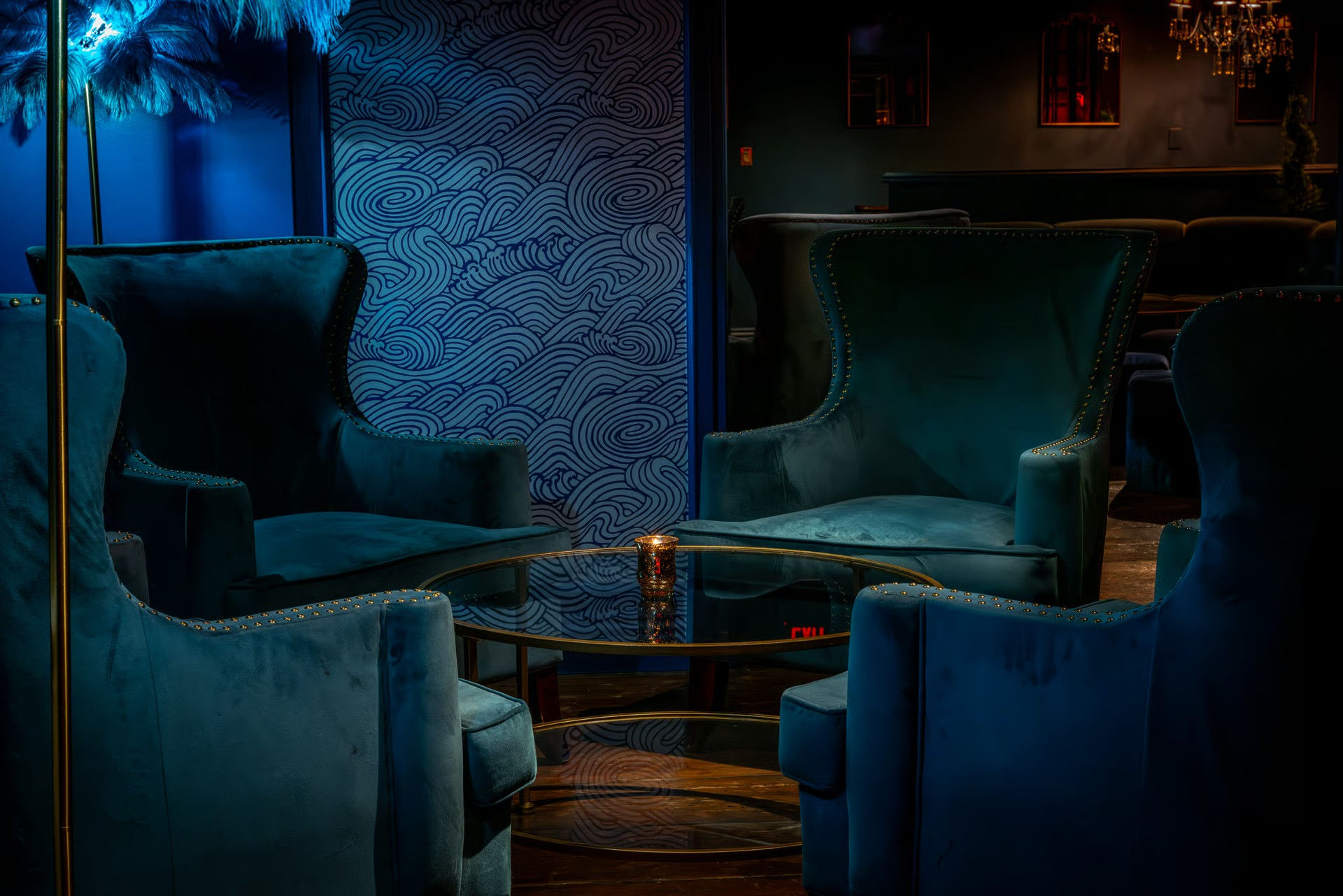 Intimate cocktail lounge interior with plush teal velvet wingback chairs clustered around a glass coffee table holding a candle, moody blue lighting and swirling patterned wallpaper.