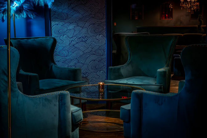 Intimate cocktail lounge interior with plush teal velvet wingback chairs clustered around a glass coffee table holding a candle, moody blue lighting and swirling patterned wallpaper.