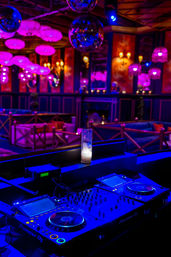 Vibrant neon-lit nightclub interior with DJ booth and professional decks/mixer, disco balls and a glowing candle above plush lounge seating bathed in purple-pink light.