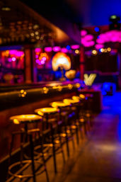 Neon-lit bar interior at night with glowing yellow bar stools, purple-pink bokeh lights and vibrant nightlife ambiance.