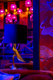 Neon-lit lounge interior featuring a gold crane table lamp with a black shade, magenta wall sconces, red crane-pattern wallpaper and plush velvet seating.