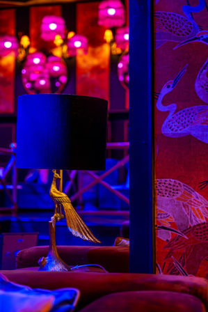 Neon-lit lounge interior featuring a gold crane table lamp with a black shade, magenta wall sconces, red crane-pattern wallpaper and plush velvet seating.