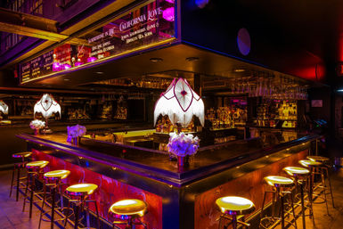 Moody neon-lit cocktail bar interior in a city nightlife setting with a U-shaped polished bar, glowing gold bar stools, vintage fringed lamps, hanging glassware, illuminated bottles and floral centerpieces.