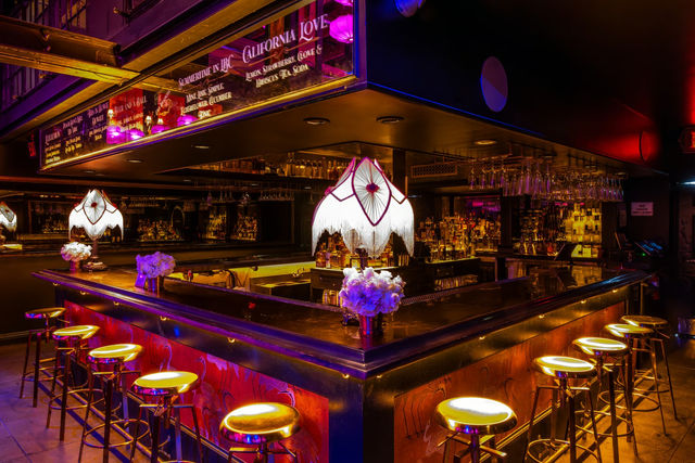Moody neon-lit cocktail bar interior in a city nightlife setting with a U-shaped polished bar, glowing gold bar stools, vintage fringed lamps, hanging glassware, illuminated bottles and floral centerpieces.