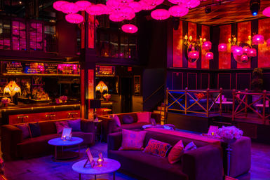Vibrant urban lounge bar interior at night with pink hanging lanterns, purple neon glow, plush velvet sofas, low marble tables, floral accents and an illuminated cocktail bar for cozy VIP seating.