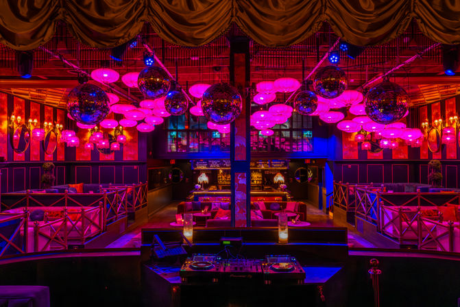 Upscale nightclub lounge interior with DJ booth in foreground, plush VIP seating and cocktail bar beneath mirrored disco balls and magenta-pink lantern lighting