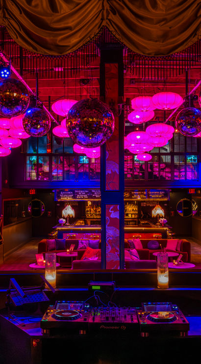 Upscale nightclub lounge interior with DJ booth in foreground, plush VIP seating and cocktail bar beneath mirrored disco balls and magenta-pink lantern lighting