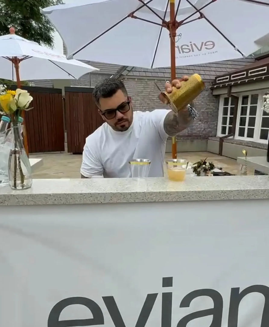 Man in a white tee and sunglasses pouring a shaken cocktail into clear cups at an outdoor patio bar under white umbrellas, yellow roses in a vase on the counter.