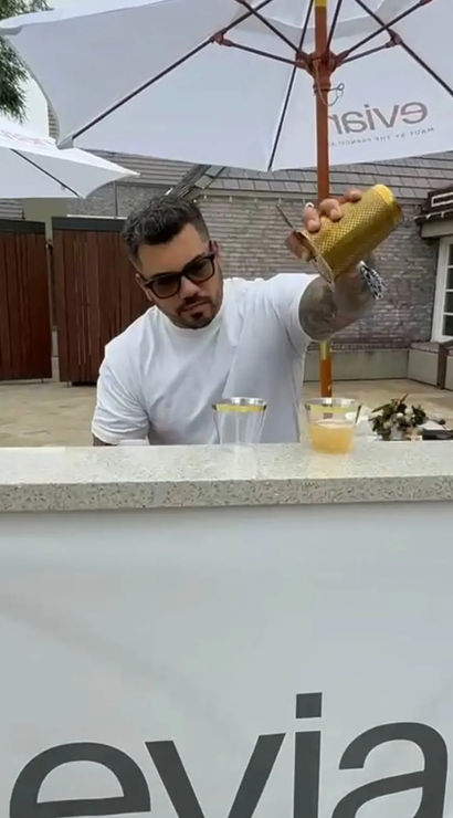 Man in a white tee and sunglasses pouring a shaken cocktail into clear cups at an outdoor patio bar under white umbrellas, yellow roses in a vase on the counter.