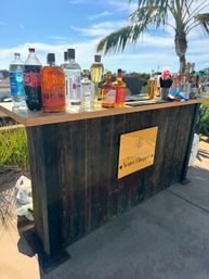 Sunny outdoor wooden bar on a patio with assorted liquor bottles, mixers and bar tools on the counter, a yellow champagne sign on the front and a palm tree with blue sky in the background.