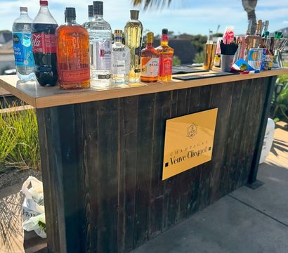 Sunny outdoor wooden bar on a patio with assorted liquor bottles, mixers and bar tools on the counter, a yellow champagne sign on the front and a palm tree with blue sky in the background.