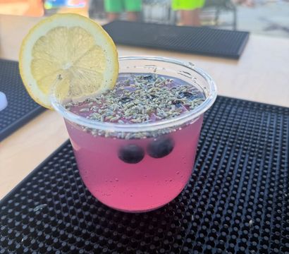 Refreshing pink lavender lemonade in a clear plastic cup, topped with lavender buds, blueberries and a lemon wheel, set on a black bar mat at a poolside outdoor bar.