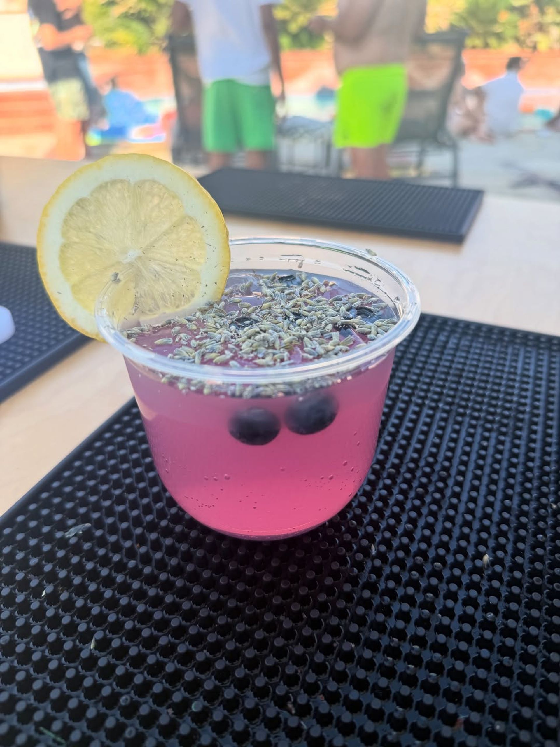 Refreshing pink lavender lemonade in a clear plastic cup, topped with lavender buds, blueberries and a lemon wheel, set on a black bar mat at a poolside outdoor bar.