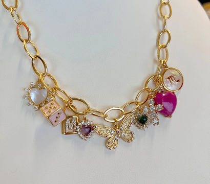 Close-up of a gold charm necklace on a white display with a filigree butterfly centerpiece, pink enamel and heart-shaped gemstone charms, mother-of-pearl disc and sparkling crystal accents — women's fashion jewelry.