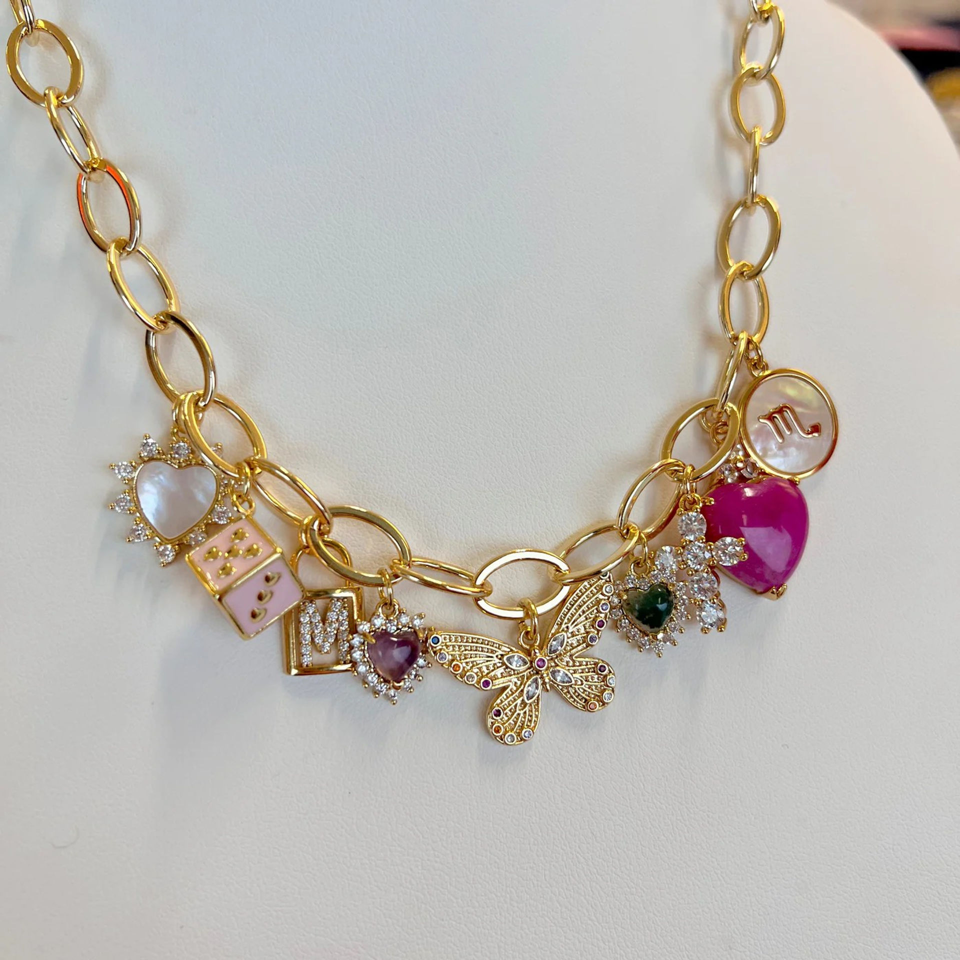 Close-up of a gold charm necklace on a white display with a filigree butterfly centerpiece, pink enamel and heart-shaped gemstone charms, mother-of-pearl disc and sparkling crystal accents — women's fashion jewelry.