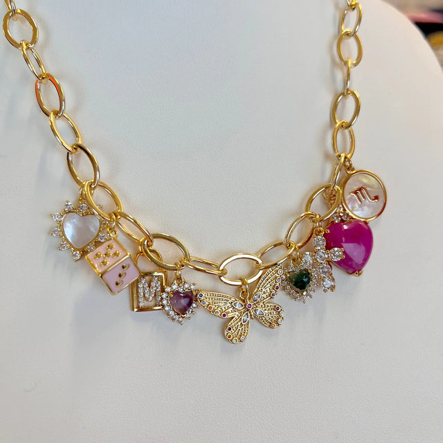 Close-up of a gold charm necklace on a white display with a filigree butterfly centerpiece, pink enamel and heart-shaped gemstone charms, mother-of-pearl disc and sparkling crystal accents — women's fashion jewelry.