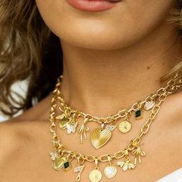 Close-up of a model's neck wearing layered chunky gold charm necklaces — hearts, bows, star coins, a green gemstone and a '7' charm, stylish women's gold jewelry detail.