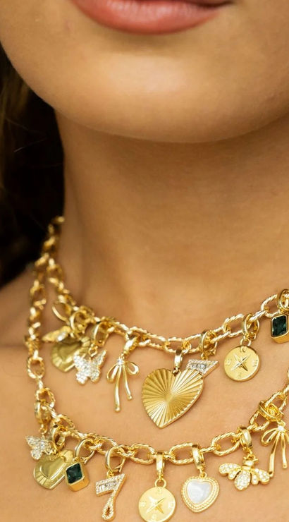 Close-up of a model's neck wearing layered chunky gold charm necklaces — hearts, bows, star coins, a green gemstone and a '7' charm, stylish women's gold jewelry detail.