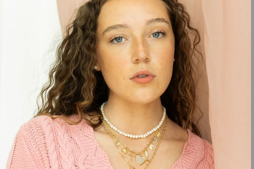 Chic close-up portrait of a freckled young woman with curly brown hair and blue eyes, wearing layered pearl and gold necklaces and a pink knit sweater against a soft pink backdrop.