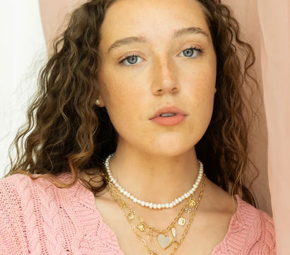 Chic close-up portrait of a freckled young woman with curly brown hair and blue eyes, wearing layered pearl and gold necklaces and a pink knit sweater against a soft pink backdrop.