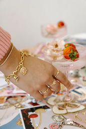 Close-up of a hand wearing a gold charm bracelet and delicate rings reaching for a pink coupe glass garnished with strawberries and a pale flower on a pastel brunch tablescape.