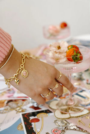 Close-up of a hand wearing a gold charm bracelet and delicate rings reaching for a pink coupe glass garnished with strawberries and a pale flower on a pastel brunch tablescape.